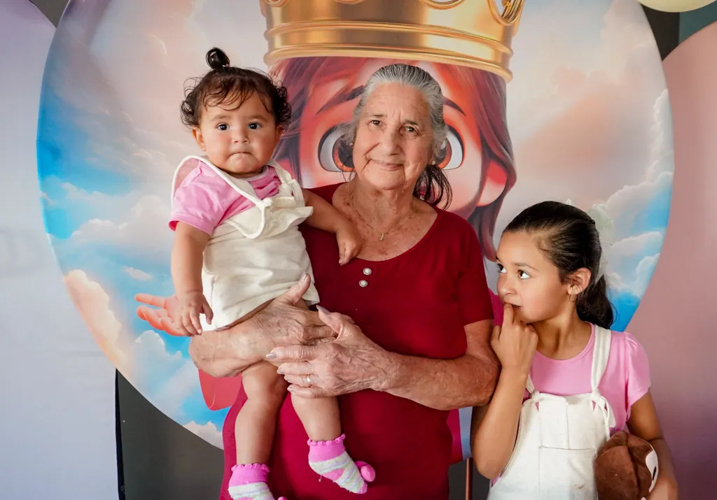 Senhora segurando um bebê ao lado de uma menina, em frente a um painel de festa