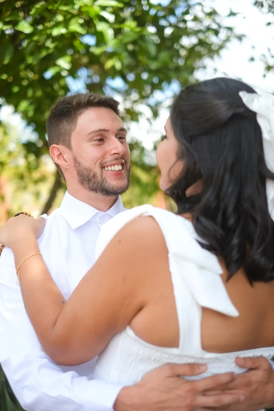 Noivos abraçados, trocando olhares sorridentes sob as árvores