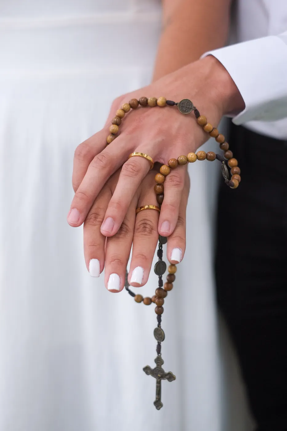 Mãos dos noivos sobrepostas, com as alianças douradas e um terço de madeira