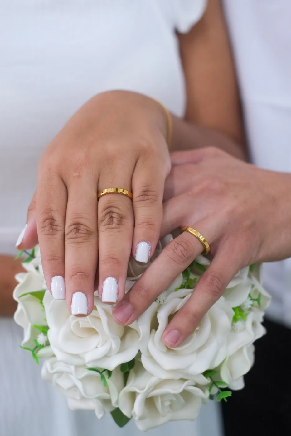 Mãos dos noivos com as alianças douradas sobre um buquê de rosas brancas