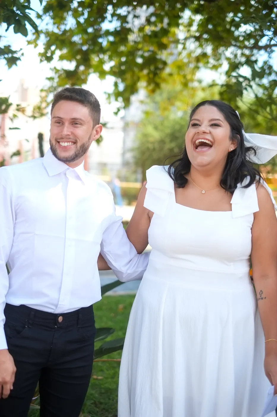 Casal de noivos rindo ao ar livre, ele de camisa branca e ela de vestido branco com véu